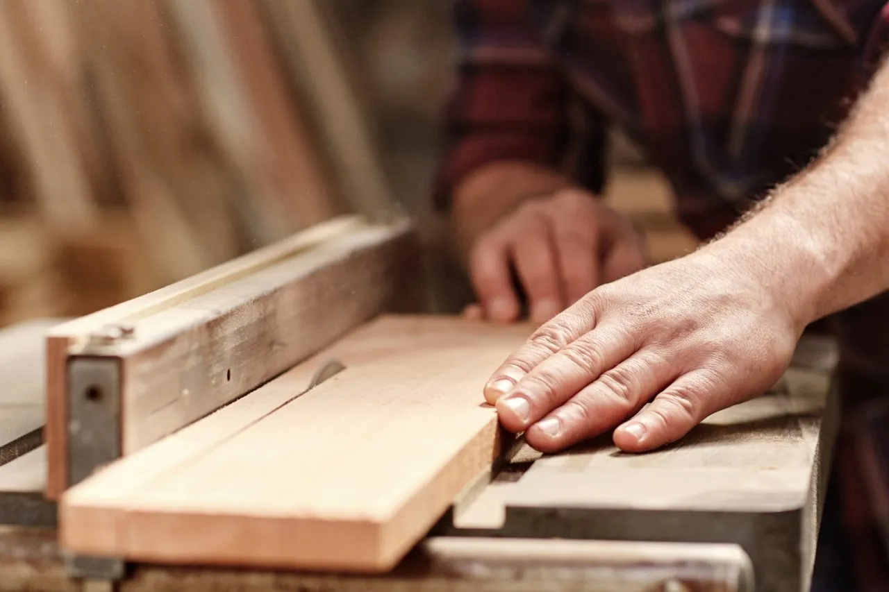 Primer plano de una mano de carpintero experto guiando una tabla de madera clara en una sierra de banco de precisión en el taller de fabricación de mobiliario de oficina de Tamasa Office Design